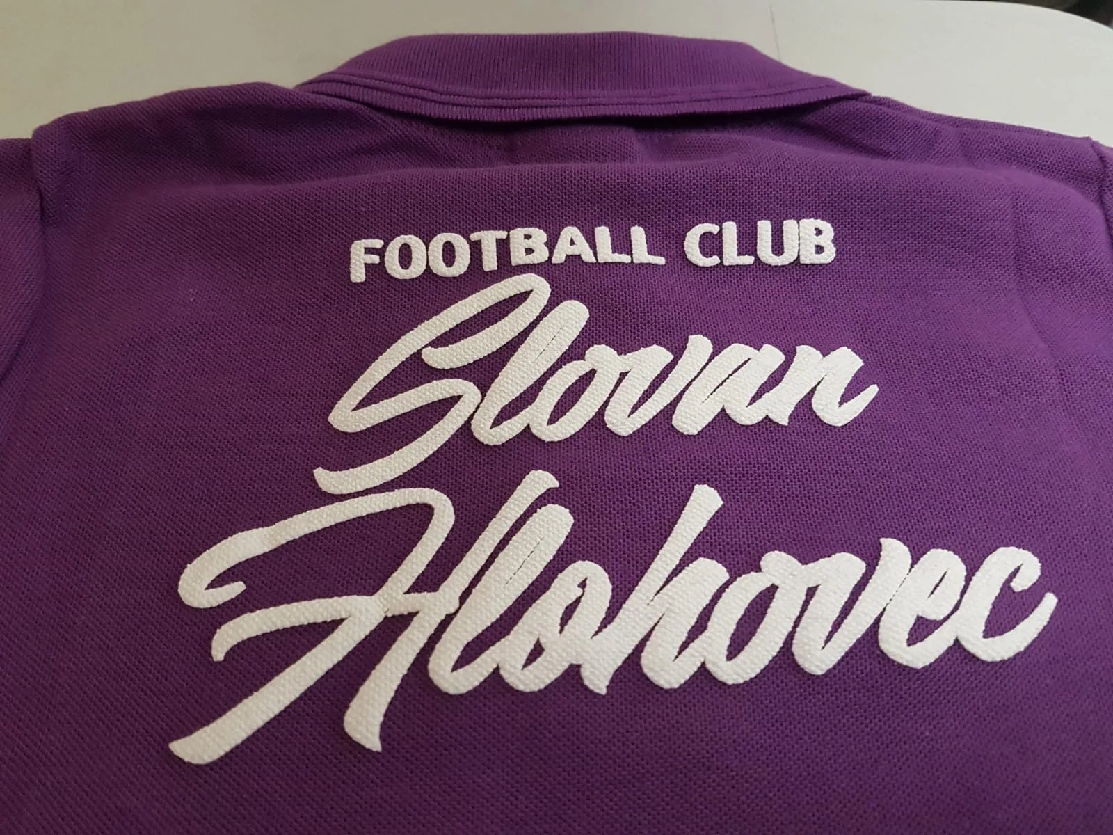 Biely vyšitý nápis futbalového klubu na fialovej polokošeli