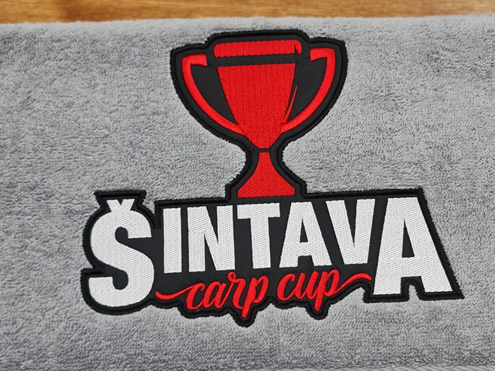 Vyšité logo rybárskej súťaže s pohárom na sivom uteráku