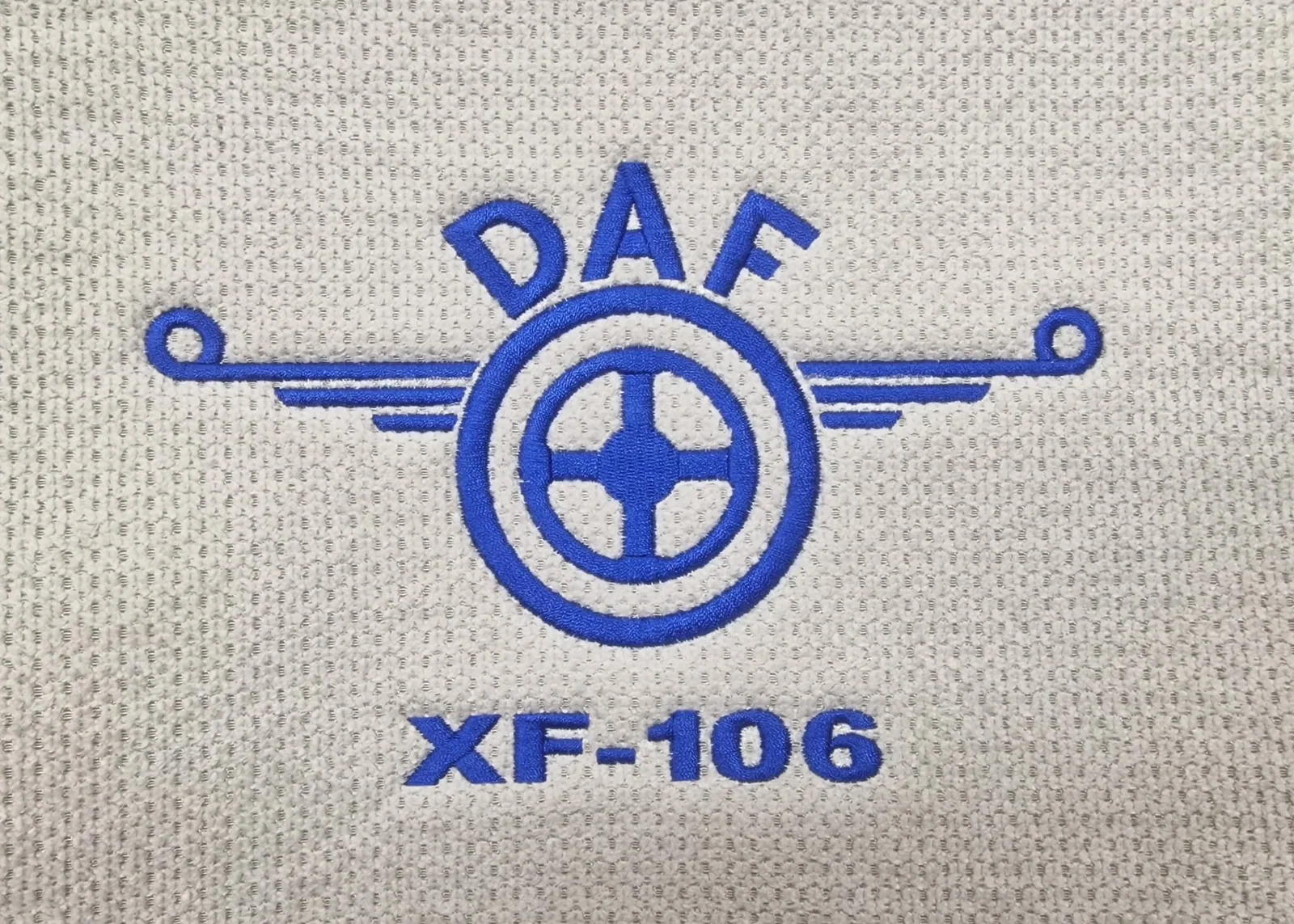 Modré vyšité logo s volantom a krídlami na sivom textile