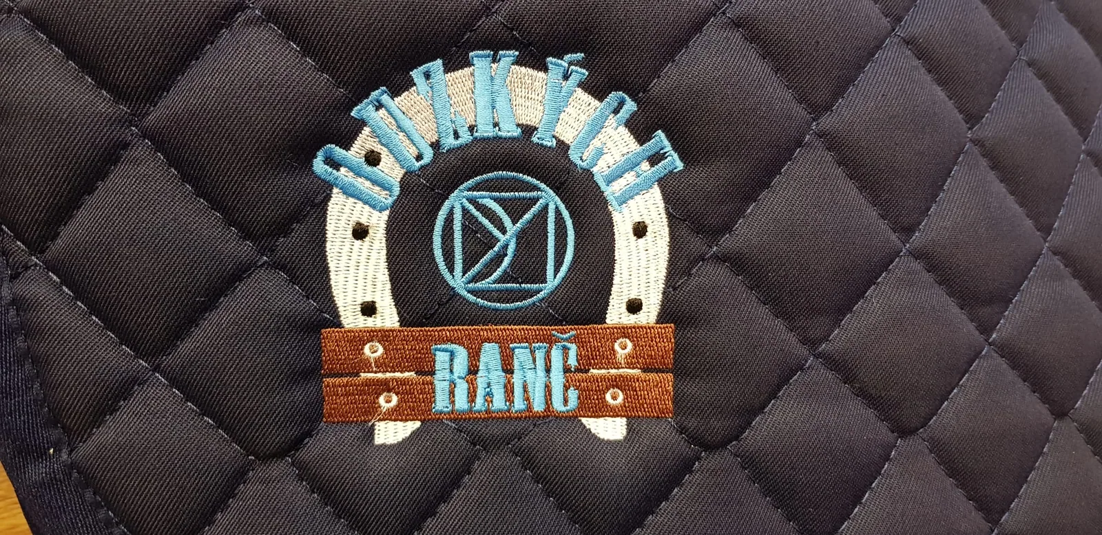 Vyšité logo ranča na hnedej prešívanej podsedlovej deke